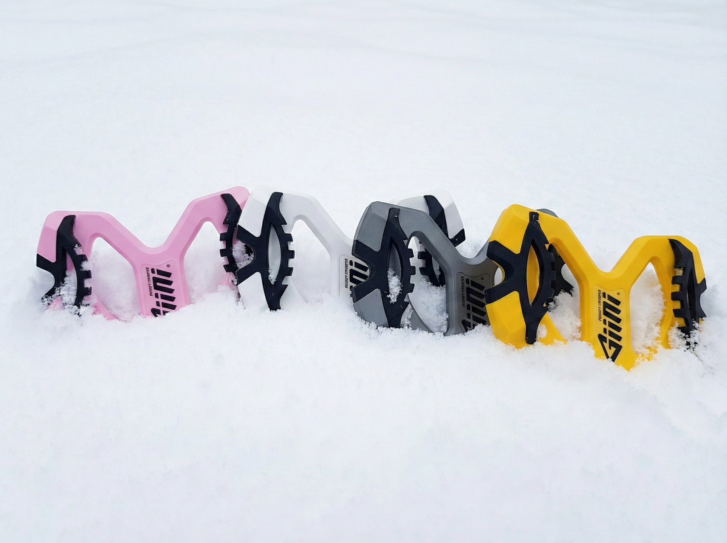 A close-up photograph shows four Giini snowboard control devices arranged in a horizontal line, partially buried in deep white powder snow. From left to right, the devices are pink, white, black, and yellow, each with black rubber grips and the "Giini" logo.
