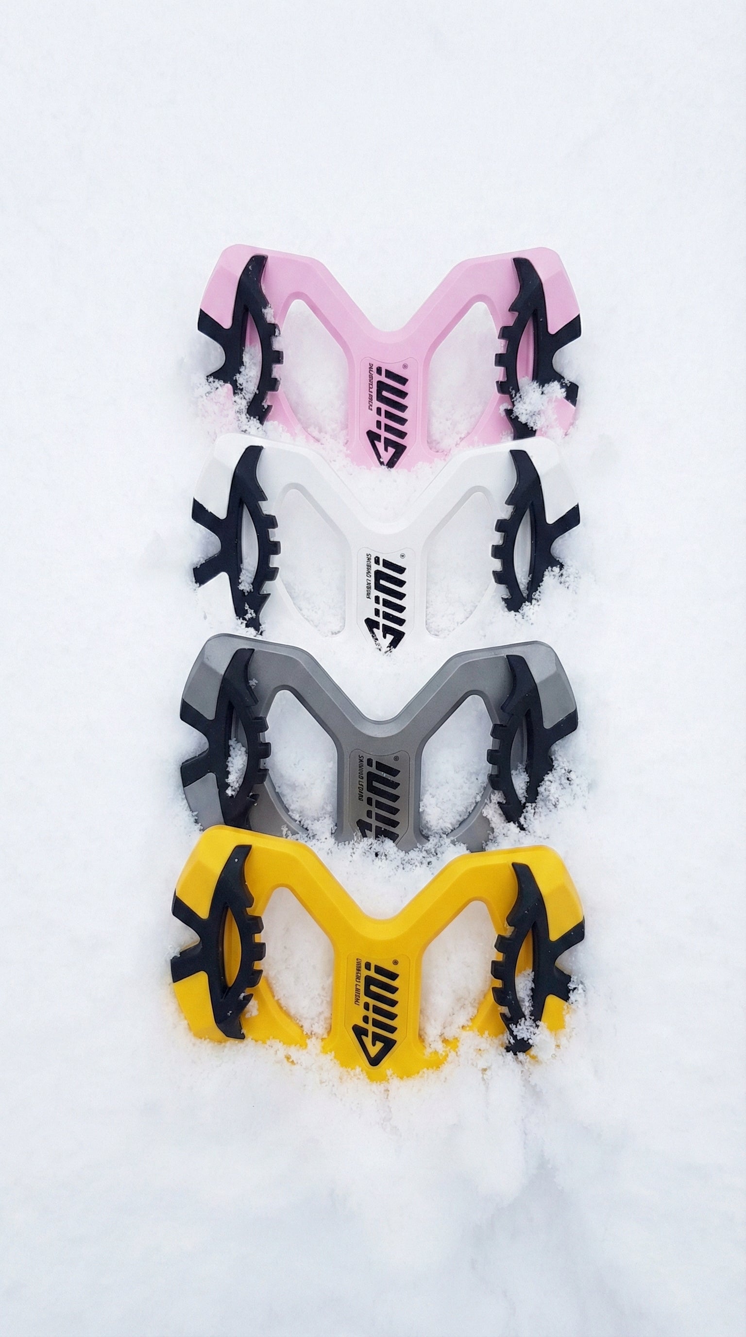 A top-down photograph shows four Giini snowboard control devices arranged in a vertical column, partially buried in fresh white snow. From top to bottom, the devices are colored pink, white, grey, and yellow, each featuring black rubber grips and the "Giini" logo.