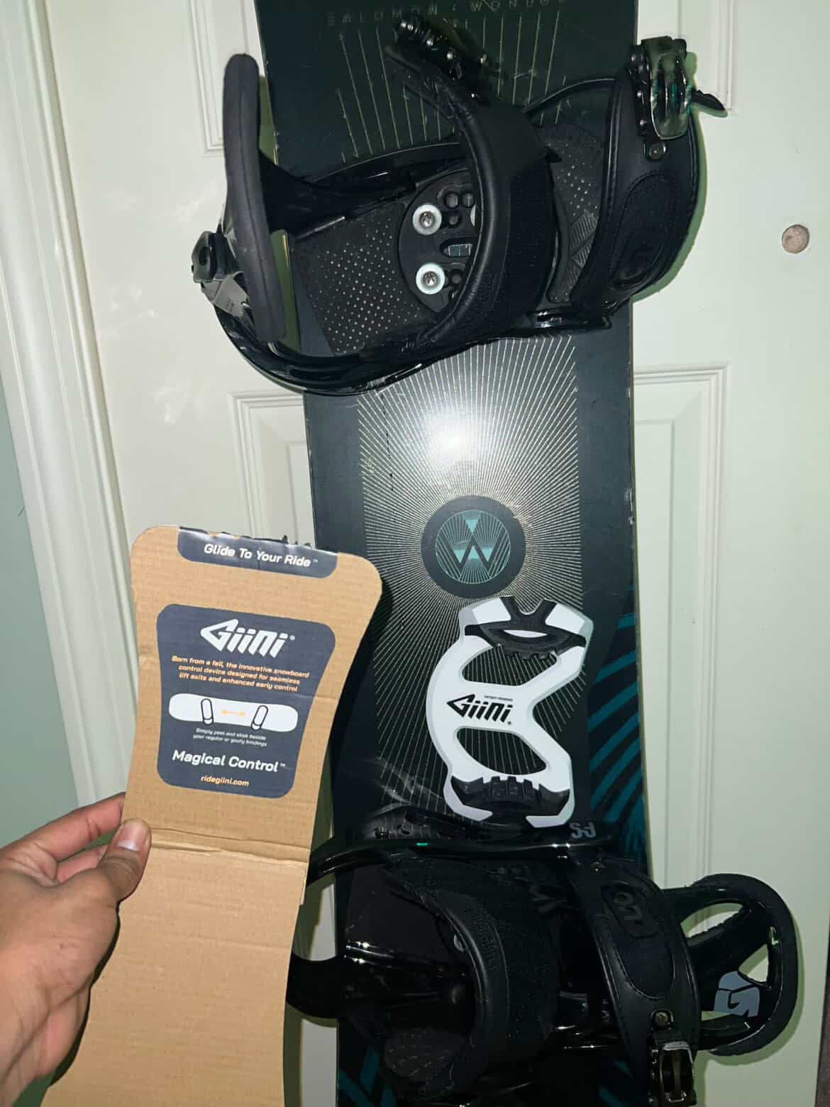 Black snowboard leaning against a door with a Giini Pyro Whiteout installed, hand holding Giini packaging