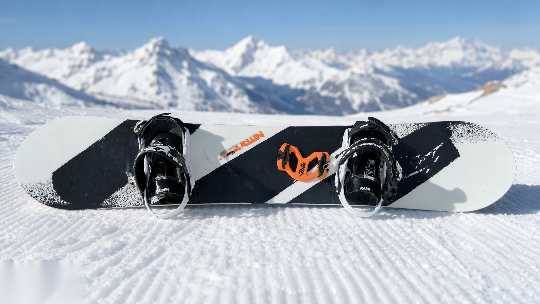 Snowboard on corduroy snow with a Giini Pyro Blaze Orange mounted between the bindings, snowy peaks behind