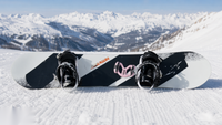 Snowboard lying on groomed snow with a Giini Pyro Powder Pink mounted between the bindings, snowy peaks behind