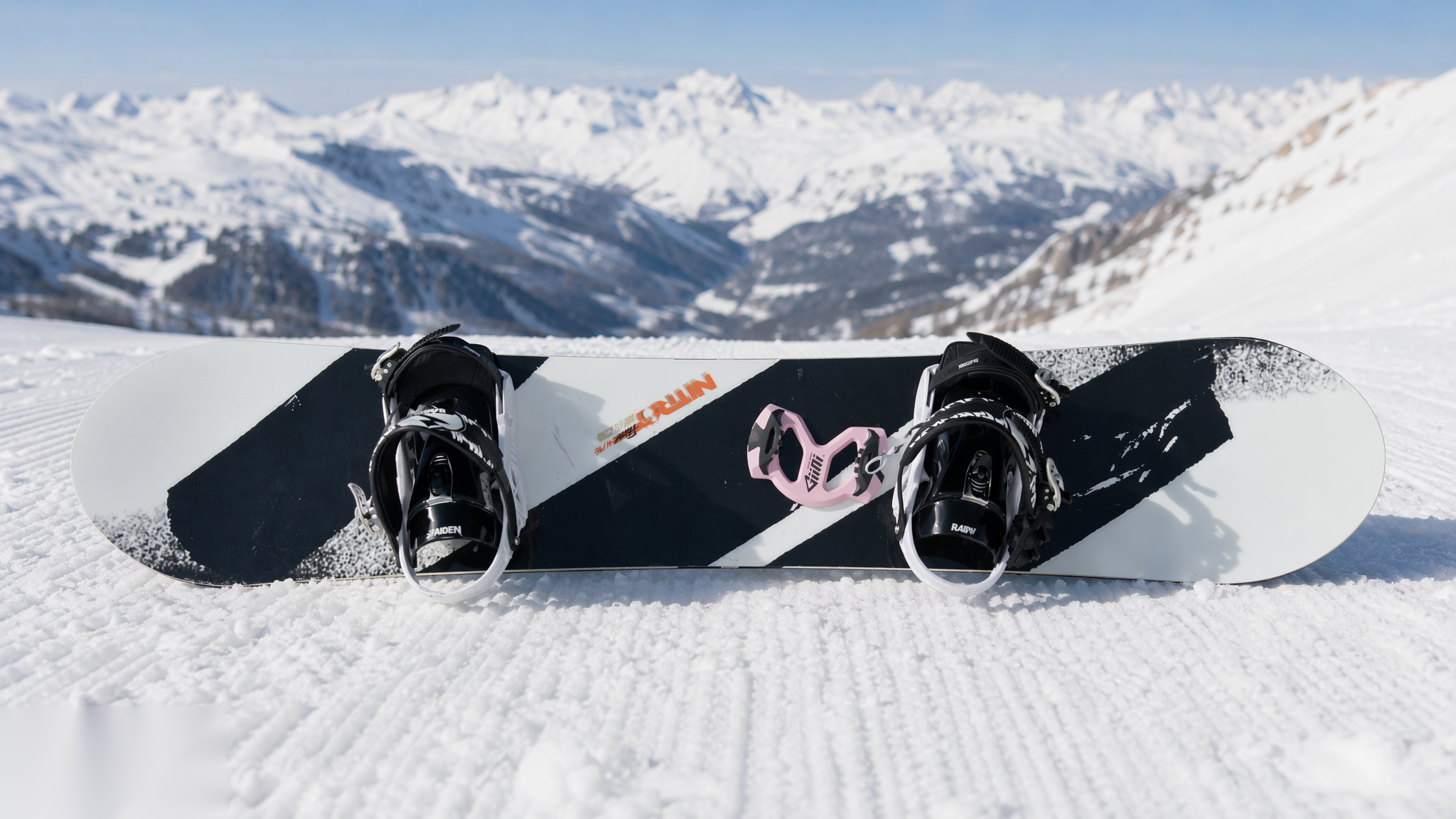 Snowboard lying on groomed snow with a Giini Pyro Powder Pink mounted between the bindings, snowy peaks behind